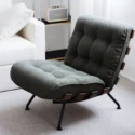A modern lounge chair with dark green tufted upholstery, black legs, and wooden accents sits next to a white sofa and a shelf holding a beige Marshall speaker in a bright, carpeted room.