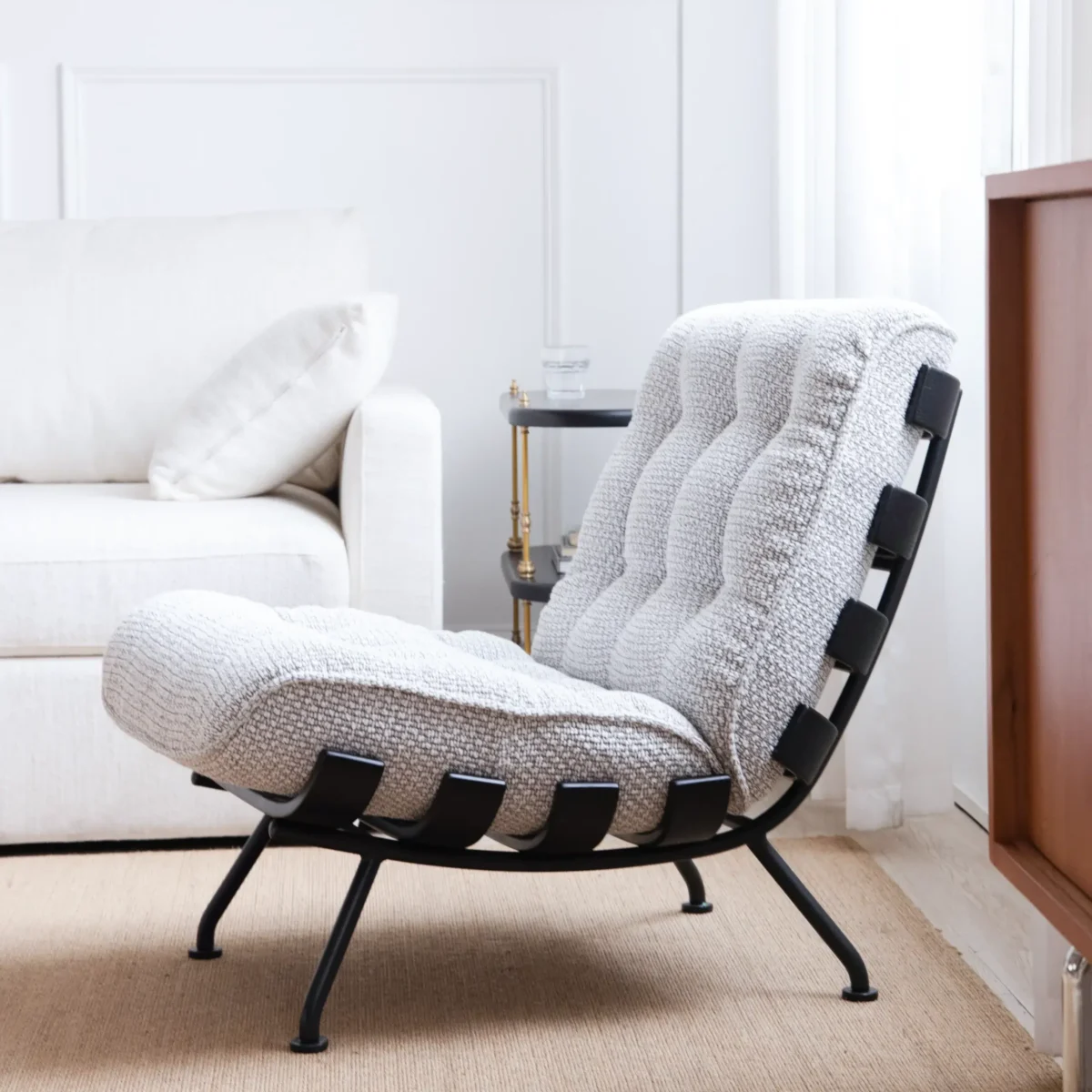 Costela Chair_Side View A modern lounge chair with a tufted, textured white cushion and black metal frame sits on a tan rug in a bright, minimalist living room next to a white sofa and a small side table.