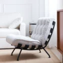 A modern lounge chair with a tufted, textured white cushion and black metal frame sits on a tan rug in a bright, minimalist living room next to a white sofa and a small side table.