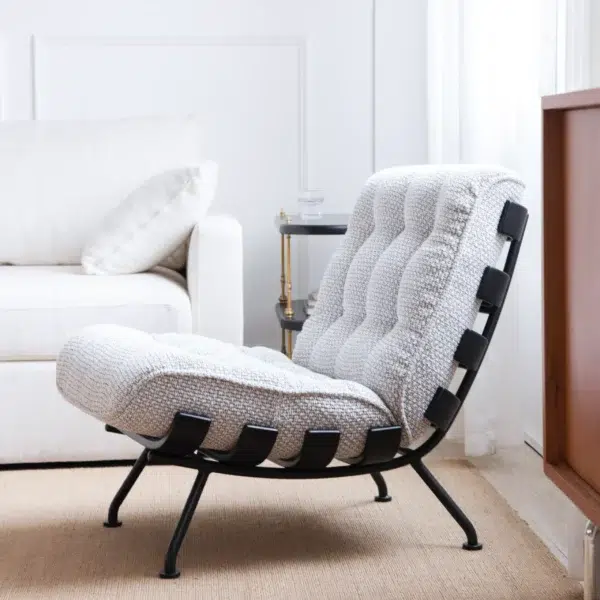 A modern lounge chair with a tufted, textured white cushion and black metal frame sits on a tan rug in a bright, minimalist living room next to a white sofa and a small side table.