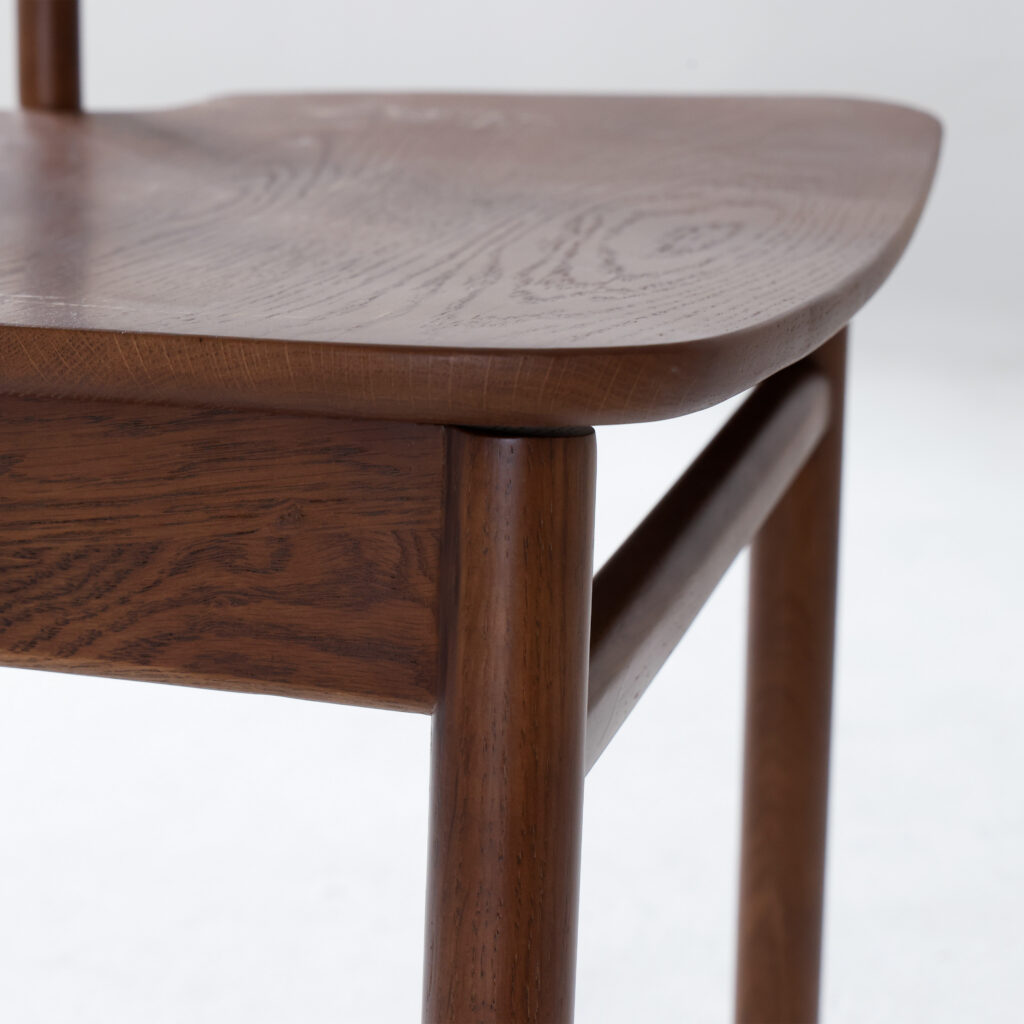 Close-up of the corner of a wooden chair with smooth, rounded edges and visible wood grain, showcasing the craftsmanship and joinery of the seat and legs against a plain white background.