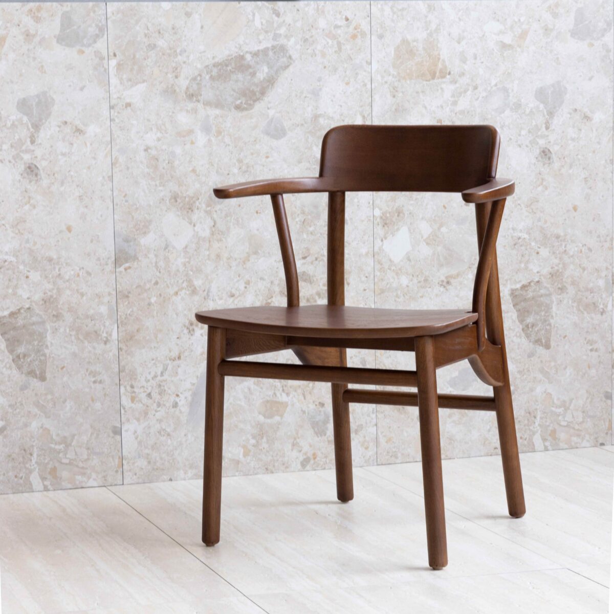 Crown Chair_Dark Brown_C1 A wooden chair with armrests and a curved backrest sits on a light tiled floor against a beige stone wall. The chair has a minimalist, mid-century modern design.