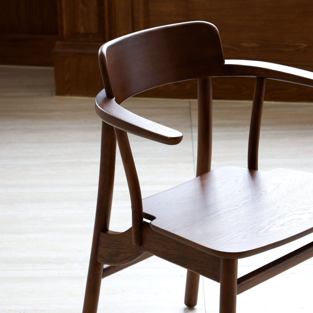 A modern wooden chair with curved armrests and a contoured backrest sits on a light-coloured floor, with wood panelling visible in the background.
