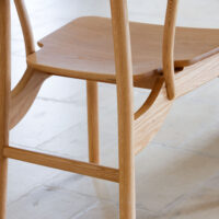 Close-up of a light wooden chair with smooth, curved lines and visible wood grain, placed on a light-coloured tiled floor.