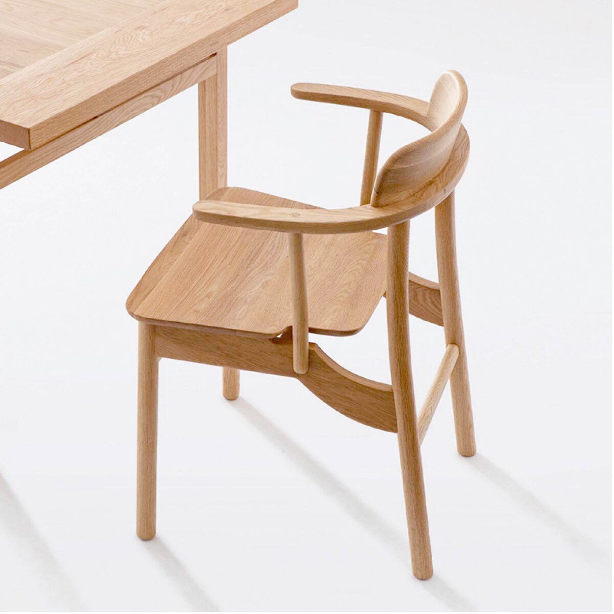 Crown Chair_Natural_C5 A light-coloured wooden chair with a curved backrest and armrests is positioned next to a matching wooden table on a white background. The furniture features a minimalist and modern design.