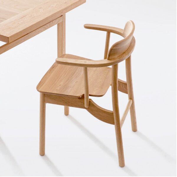 A light-coloured wooden chair with a curved backrest and armrests is positioned next to a matching wooden table on a white background. The furniture features a minimalist and modern design.