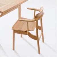 A light-coloured wooden chair with a curved backrest and armrests is positioned next to a matching wooden table on a white background. The furniture features a minimalist and modern design.