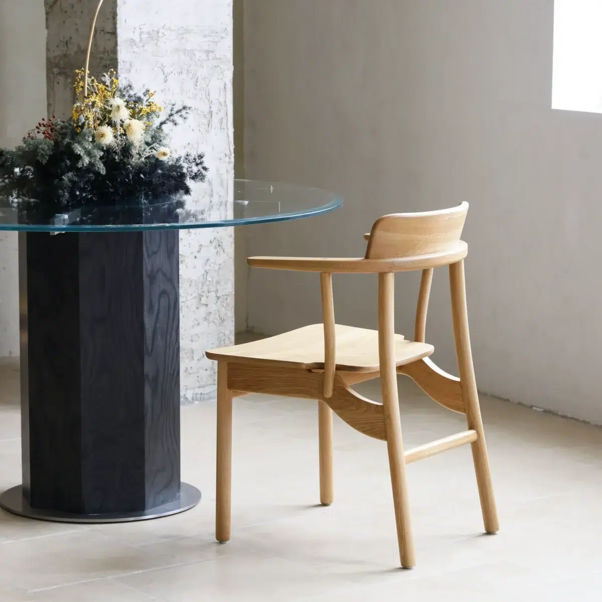 Crown Dining Chair_Back Side View A modern dining area with a round glass table on a dark cylindrical base, a light wooden chair, and a floral arrangement in a vase; sunlight streams through a window on the right.