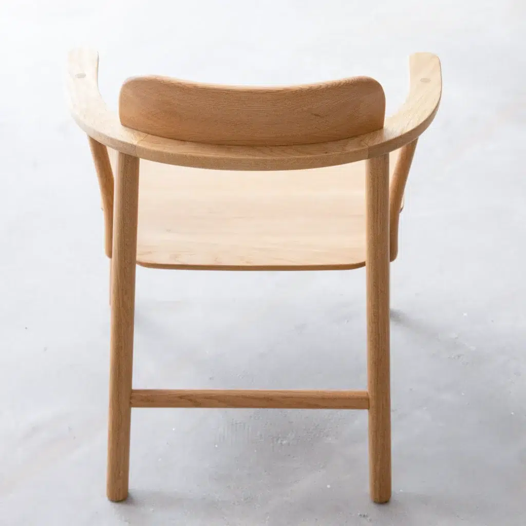 A light wooden chair with a curved backrest and armrests, viewed from behind. The chair has clean, minimalist lines and is placed on a light, textured floor.