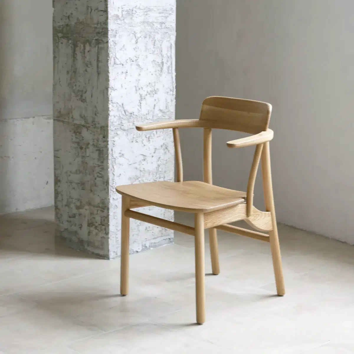 Crown Dining Chair_Front Side View A light wooden chair with a minimalist design, curved backrest, and armrests sits on a beige tiled floor next to a textured concrete pillar in a bright, modern room.