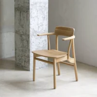 A light wooden chair with a minimalist design, curved backrest, and armrests sits on a beige tiled floor next to a textured concrete pillar in a bright, modern room.