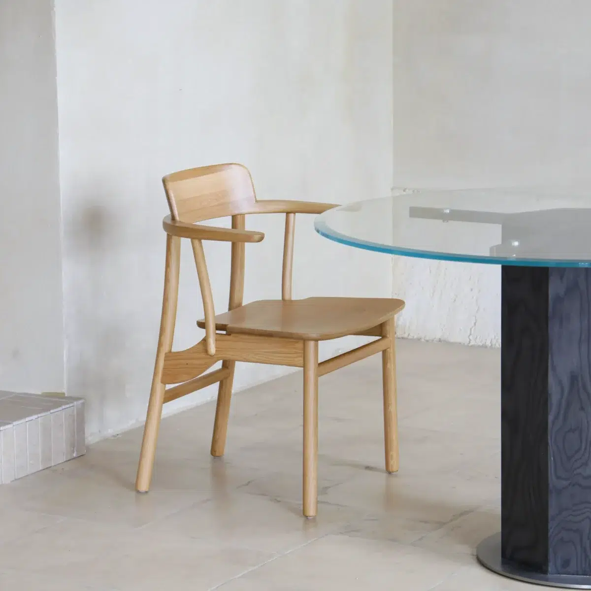 Crown Dining Chair_Front Side View(3) A wooden chair with armrests sits next to a round glass table with a black base on a beige tiled floor in a minimalist, light-coloured room.