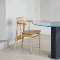 A wooden chair with armrests sits next to a round glass table with a black base on a beige tiled floor in a minimalist, light-coloured room.