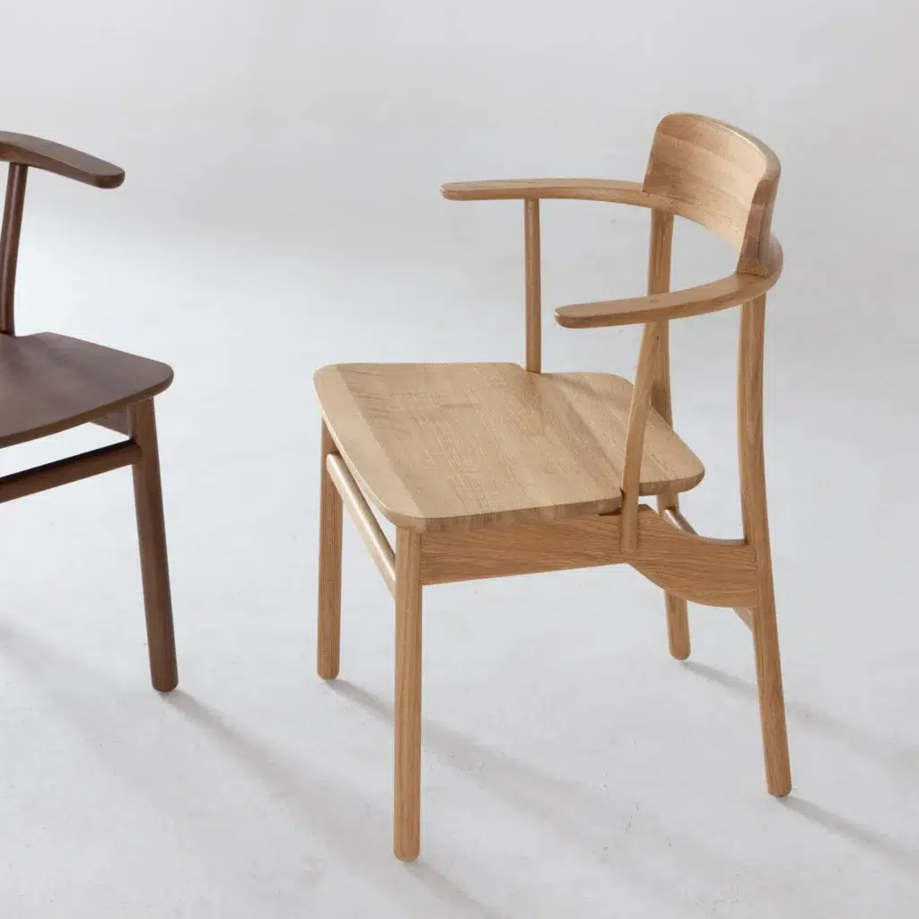 A unique wooden chair with an asymmetrical backrest and armrests, featuring a natural light wood finish. Another darker wooden chair is partially visible to the left on a white floor.