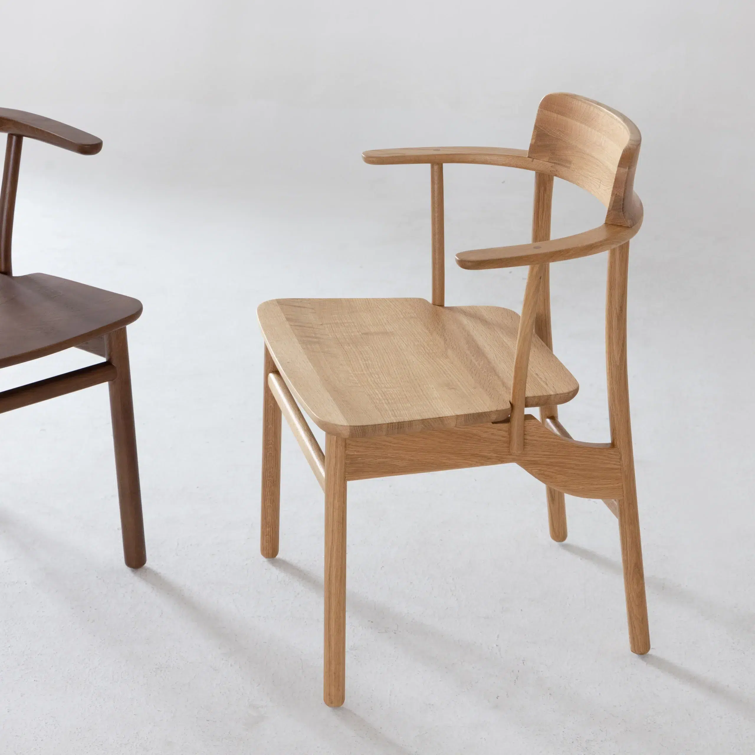 A unique wooden chair with an asymmetrical backrest and armrests, featuring a natural light wood finish. Another darker wooden chair is partially visible to the left on a white floor.