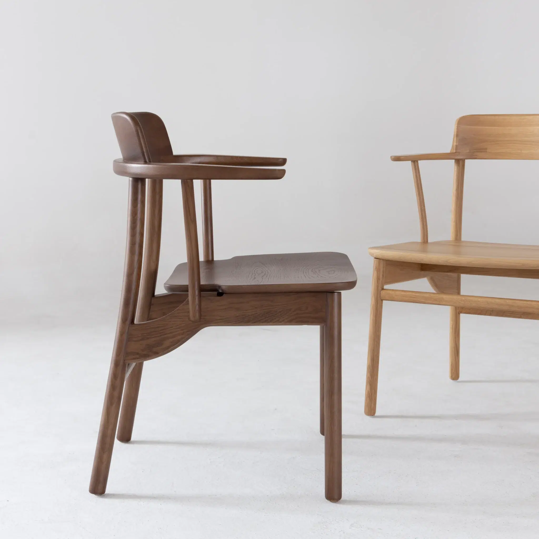 Two wooden chairs with minimalist design are placed on a light floor against a plain background. One chair is dark brown and the other is light wood, both with curved backs and armrests.
