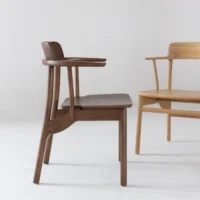 Two wooden chairs with minimalist design are placed on a light floor against a plain background. One chair is dark brown and the other is light wood, both with curved backs and armrests.