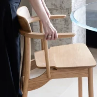 A person with a silver ring on one hand pushes a light wooden chair towards a round glass table in a modern, minimal interior.