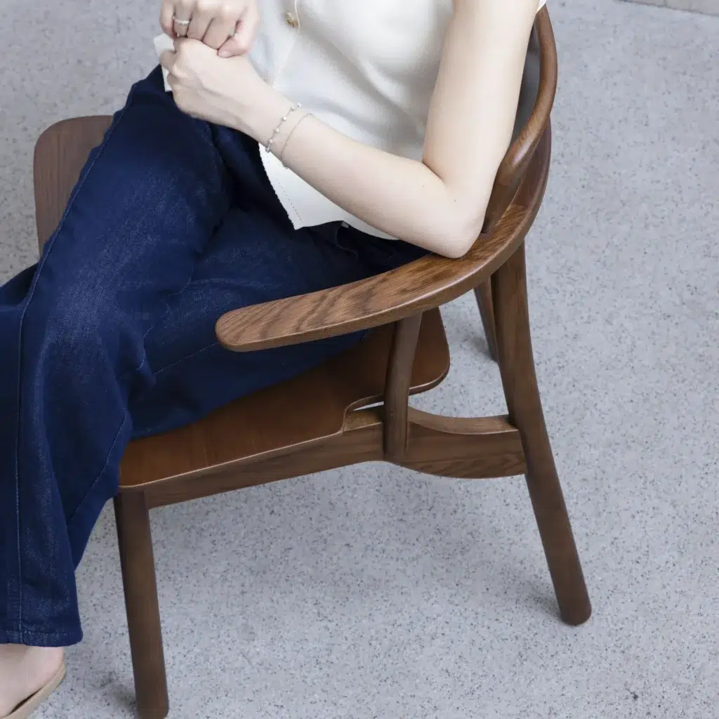 A person wearing a white sleeveless top and blue jeans sits on a wooden chair, hands clasped in their lap. The image is cropped above the shoulders and at the ankles, with a light grey floor visible.
