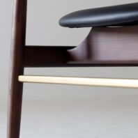 Close-up of a modern wooden chair featuring a dark wood frame, a black seat, and a horizontal brass or gold-coloured support bar near the bottom. The background is plain and light-coloured.