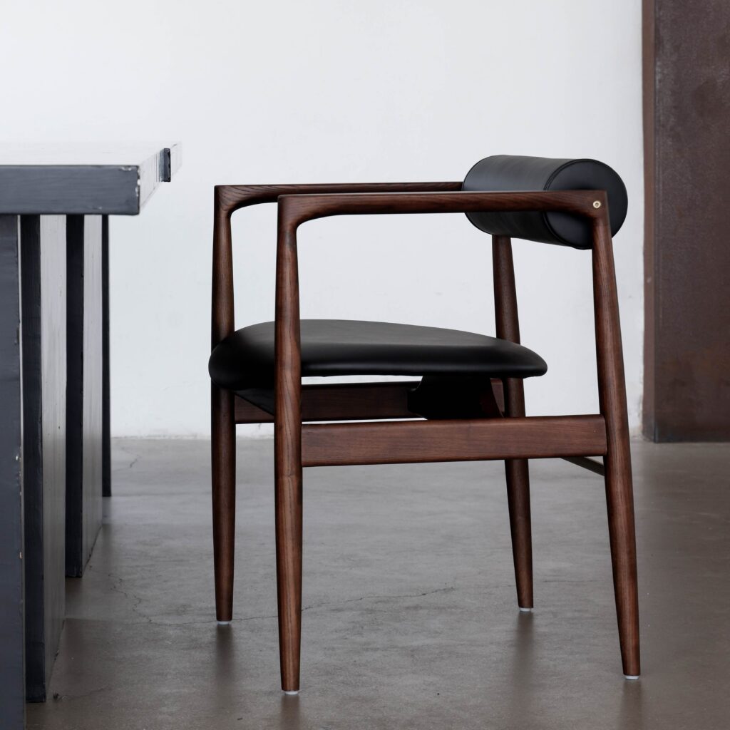 A modern wooden chair with a black leather seat and cylindrical headrest is positioned beside a dark-coloured table on a smooth concrete floor in a minimalist room.