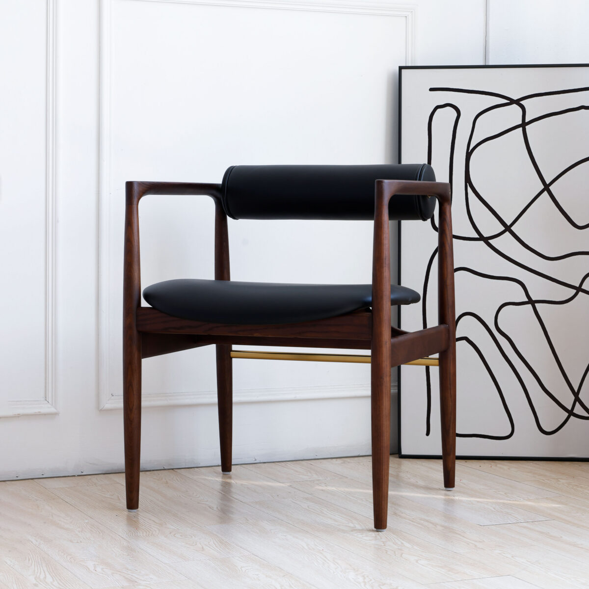 Dark Brown Frame + Black Seat_C3 A modern wooden chair with black leather seat and backrest is placed next to a wall. A large, abstract black and white artwork rests on the floor, leaning against the wall behind the chair.