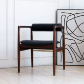 A modern wooden chair with black leather seat and backrest is placed next to a wall. A large, abstract black and white artwork rests on the floor, leaning against the wall behind the chair.