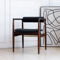 A modern wooden chair with black leather seat and backrest is placed next to a wall. A large, abstract black and white artwork rests on the floor, leaning against the wall behind the chair.