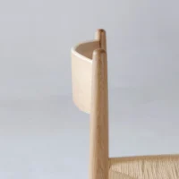 A close-up side view of a minimalist wooden chair with a curved wooden backrest and woven seat, set against a plain light background.