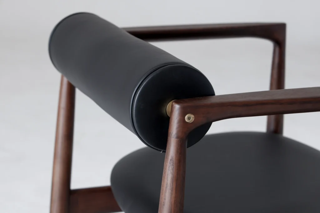 Close-up of a modern wooden chair featuring a cylindrical black leather backrest and a round black seat, with smooth, dark wood armrests and frame against a light background.