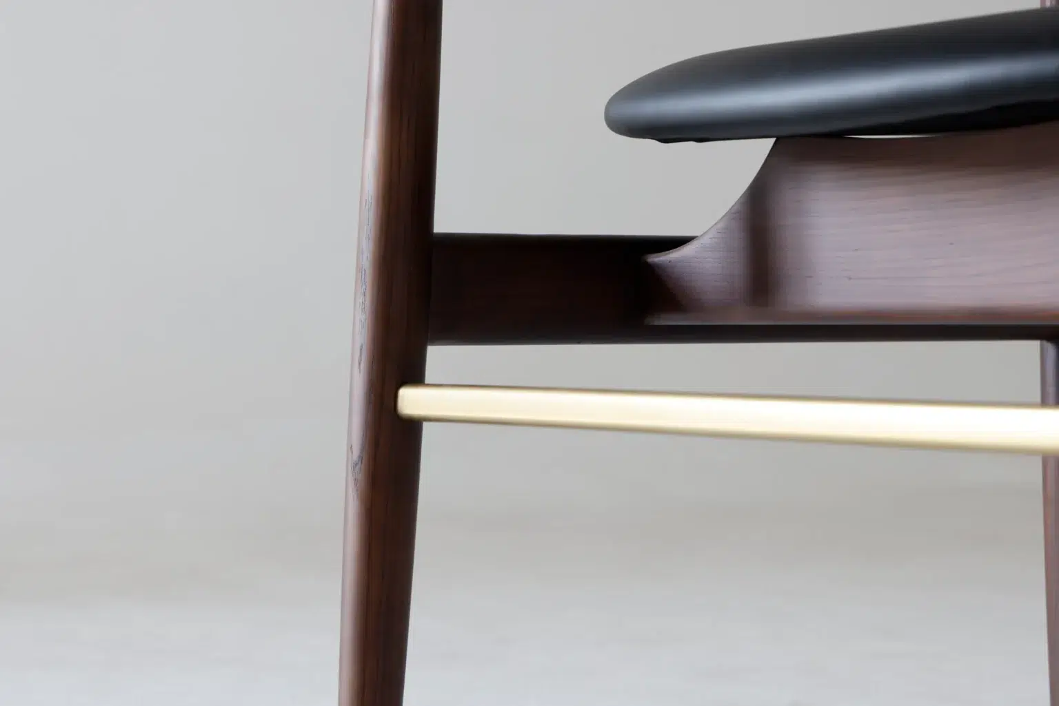Detail display2 Close-up of a modern wooden chair with a dark brown frame, black seat, and a light-coloured horizontal metal bar connecting the legs. The background is plain and out of focus.