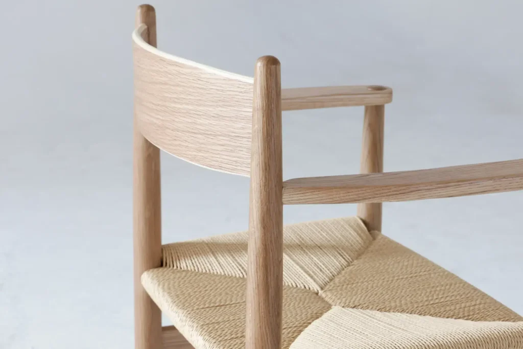 A close-up of a minimalist wooden chair with a curved backrest and woven seat, set against a plain light background. The chair features smooth, natural wood and simple, elegant lines.