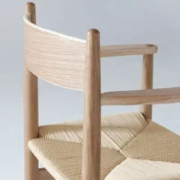 A close-up of a minimalist wooden chair with a curved backrest and woven seat, set against a plain light background. The chair features smooth, natural wood and simple, elegant lines.
