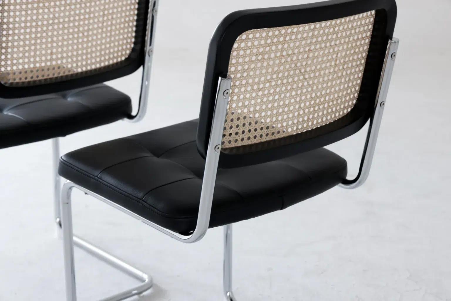 Detail display3 Two modern chairs with chrome frames, black cushioned seats, and woven cane backs are positioned on a light-coloured floor. The design is minimalist and contemporary.