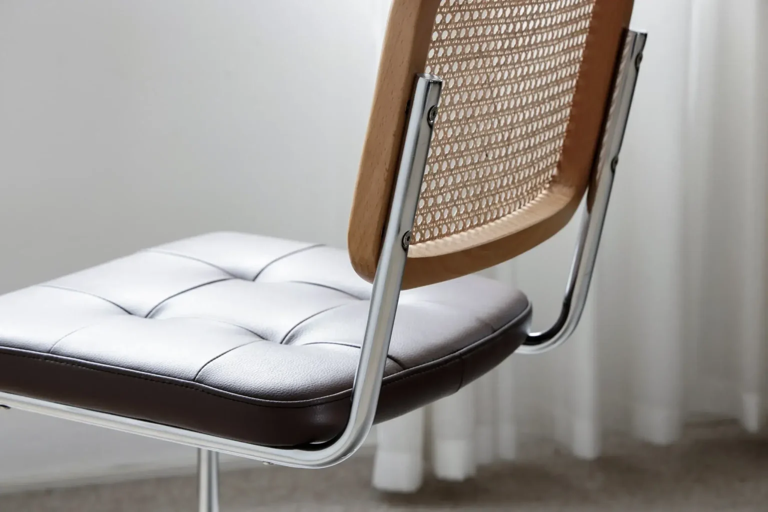 Detail display3 A close-up of a modern chair with a light, tufted leather seat, chrome legs, and a wooden backrest featuring a woven cane pattern. White curtains and a carpeted floor are visible in the background.