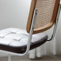 A close-up of a modern chair with a light, tufted leather seat, chrome legs, and a wooden backrest featuring a woven cane pattern. White curtains and a carpeted floor are visible in the background.