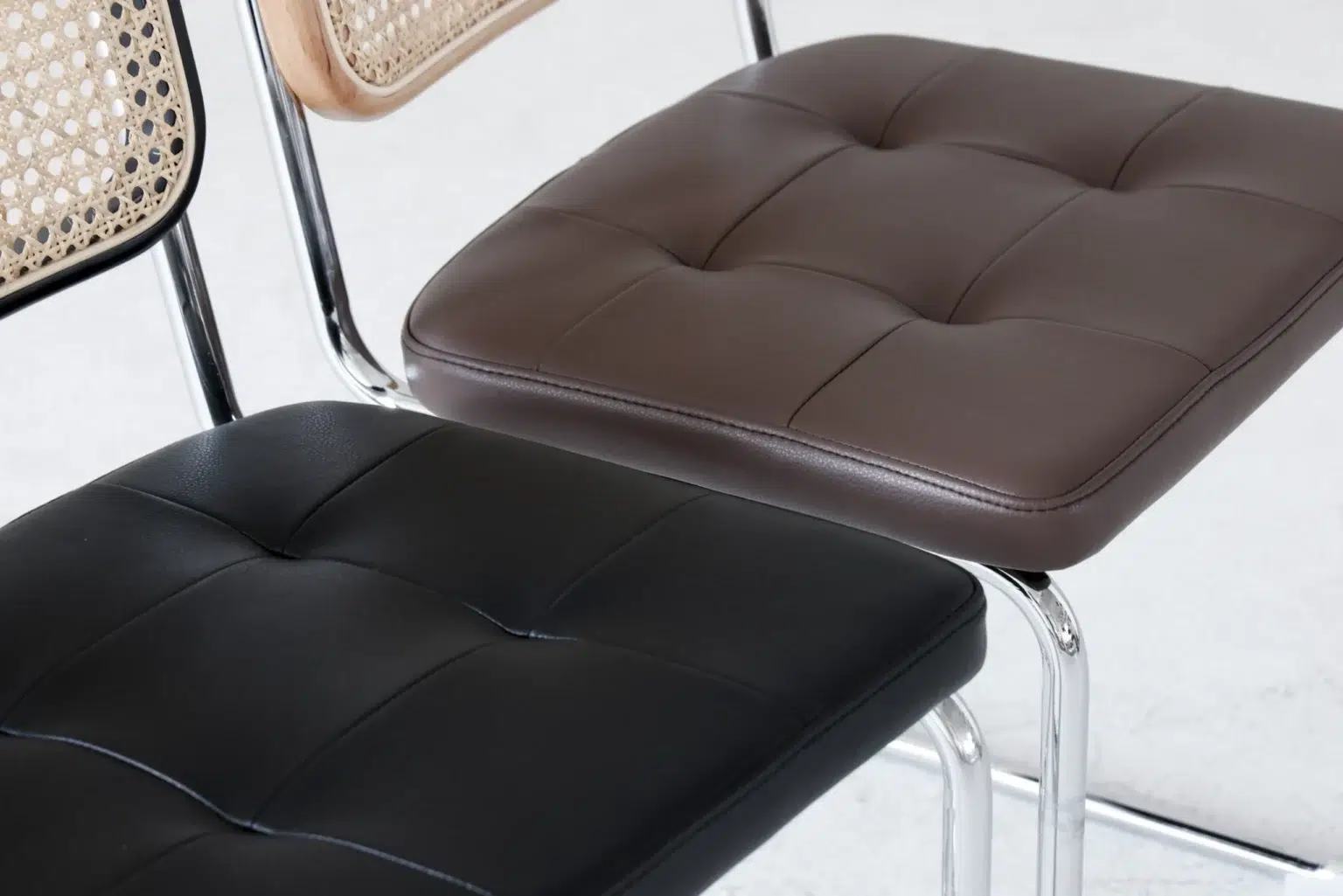 Detail display4 Close-up of two chairs with chrome legs, one with a black tufted leather cushion and the other with a brown tufted leather cushion; a third chair with a cane back is partially visible.