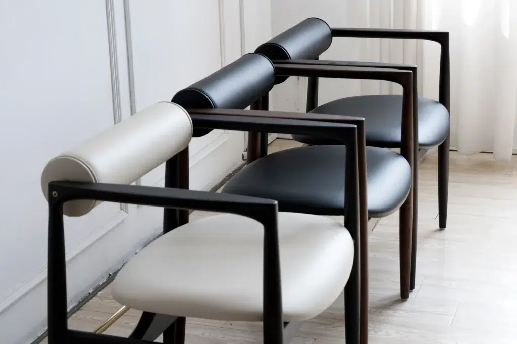 Three modern chairs in a row, each with a rounded seat and cylindrical backrest. The chairs have dark wooden frames; one is light beige, one grey, and one black. They are placed on a light wood floor near a white wall.