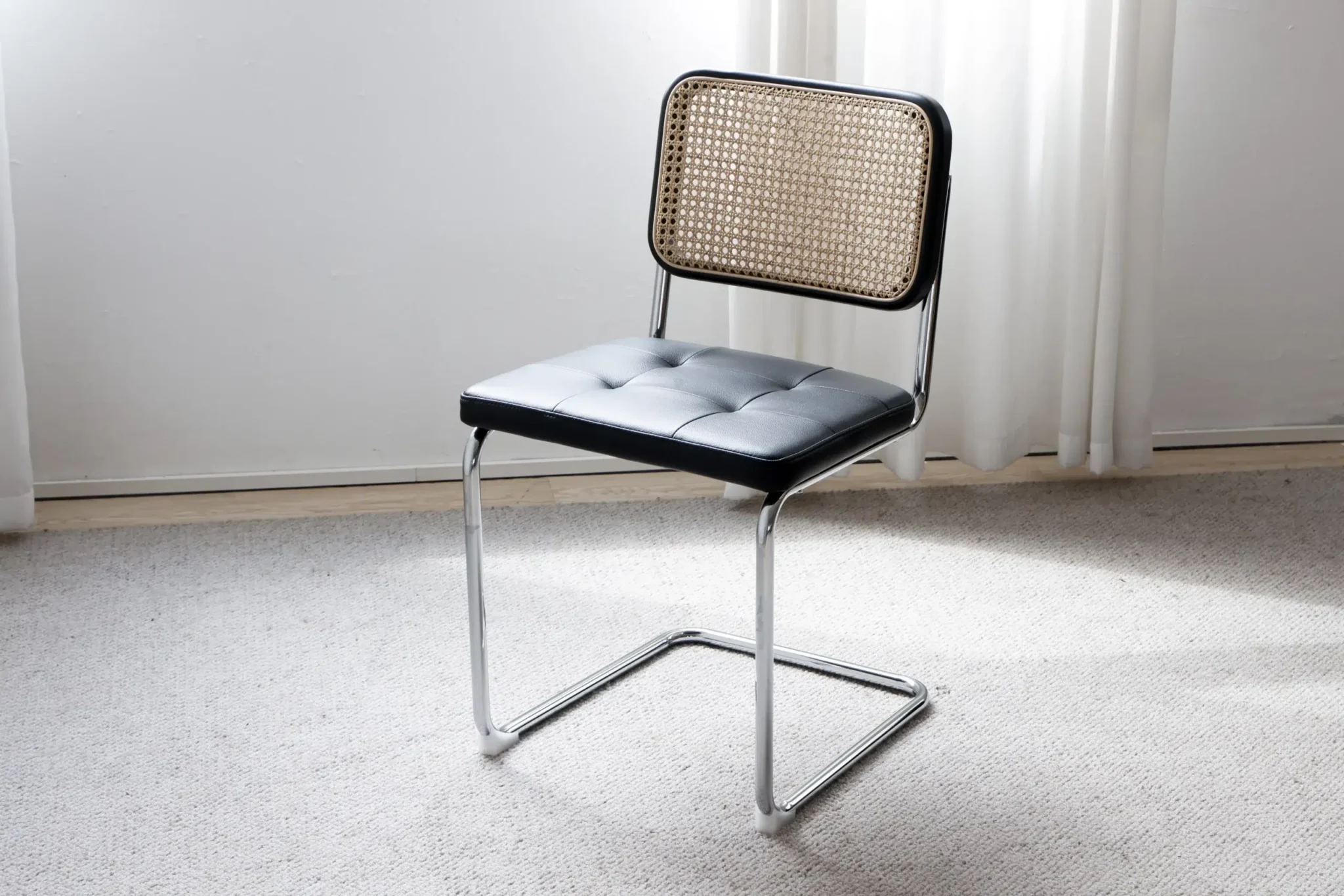 A modern chair with a black cushioned seat, a woven cane backrest, and a chrome cantilever frame sits on a light carpet near white curtains.