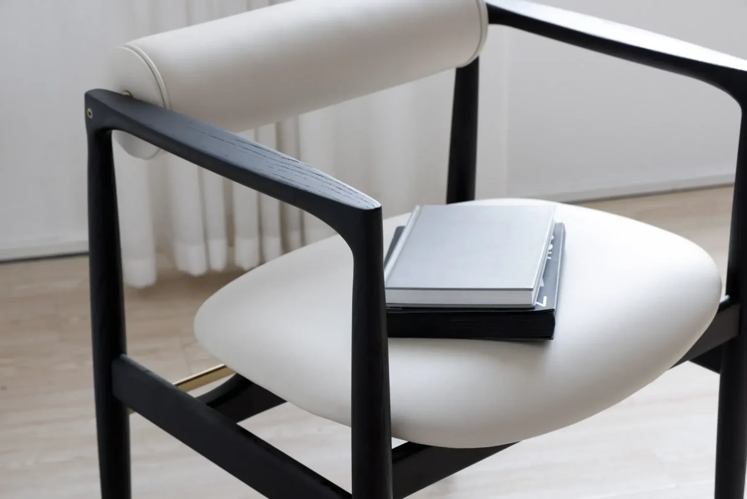 Display image1 A modern chair with a white cushioned seat and black frame, holding two closed notebooks or books stacked on top of each other, in a bright, minimalist room with light wood flooring and white curtains.