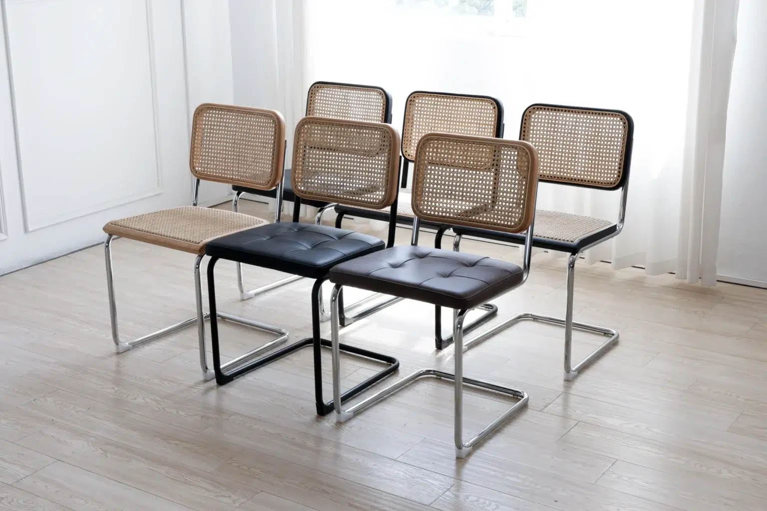 Display image2 Six modern cantilever chairs with chrome frames are arranged indoors. Four have woven cane seats and backs, while two at the front have black cushioned seats. Light wood flooring and sheer curtains are in the background.