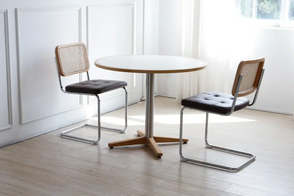 A round white table with a wooden edge is placed between two modern chairs with metal frames and cane backs in a bright, minimalist room with light wooden floors and white walls. Sunlight streams through a window.