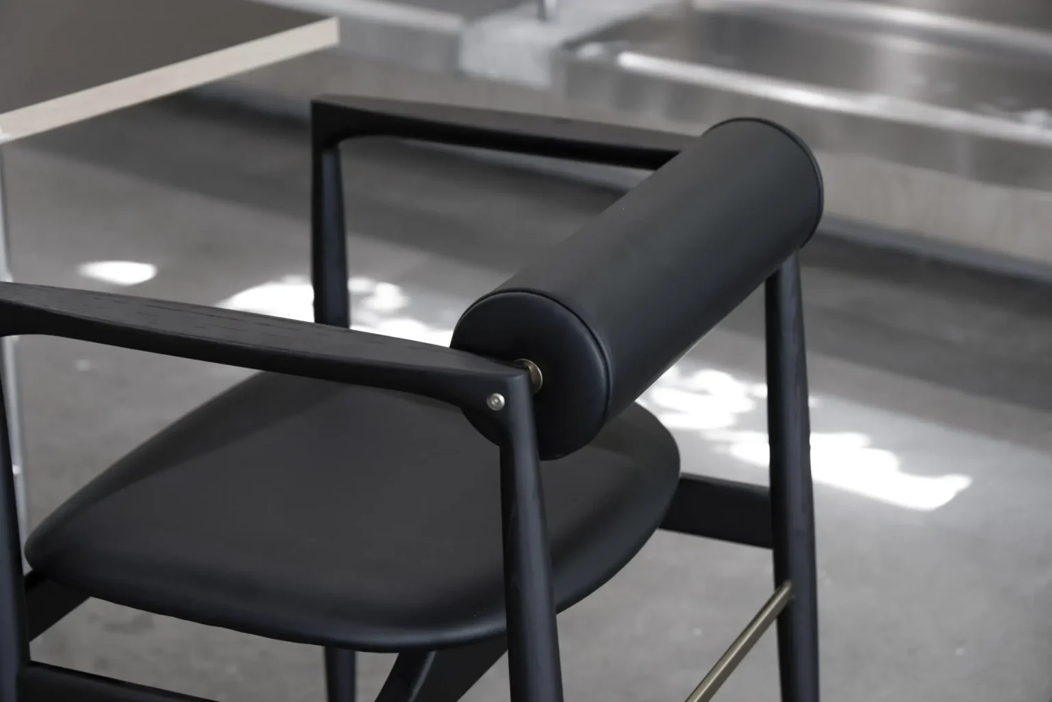Display image3 A modern black chair with a cylindrical padded backrest and sleek, angular frame is positioned next to a table in a minimalist, industrial-style interior.