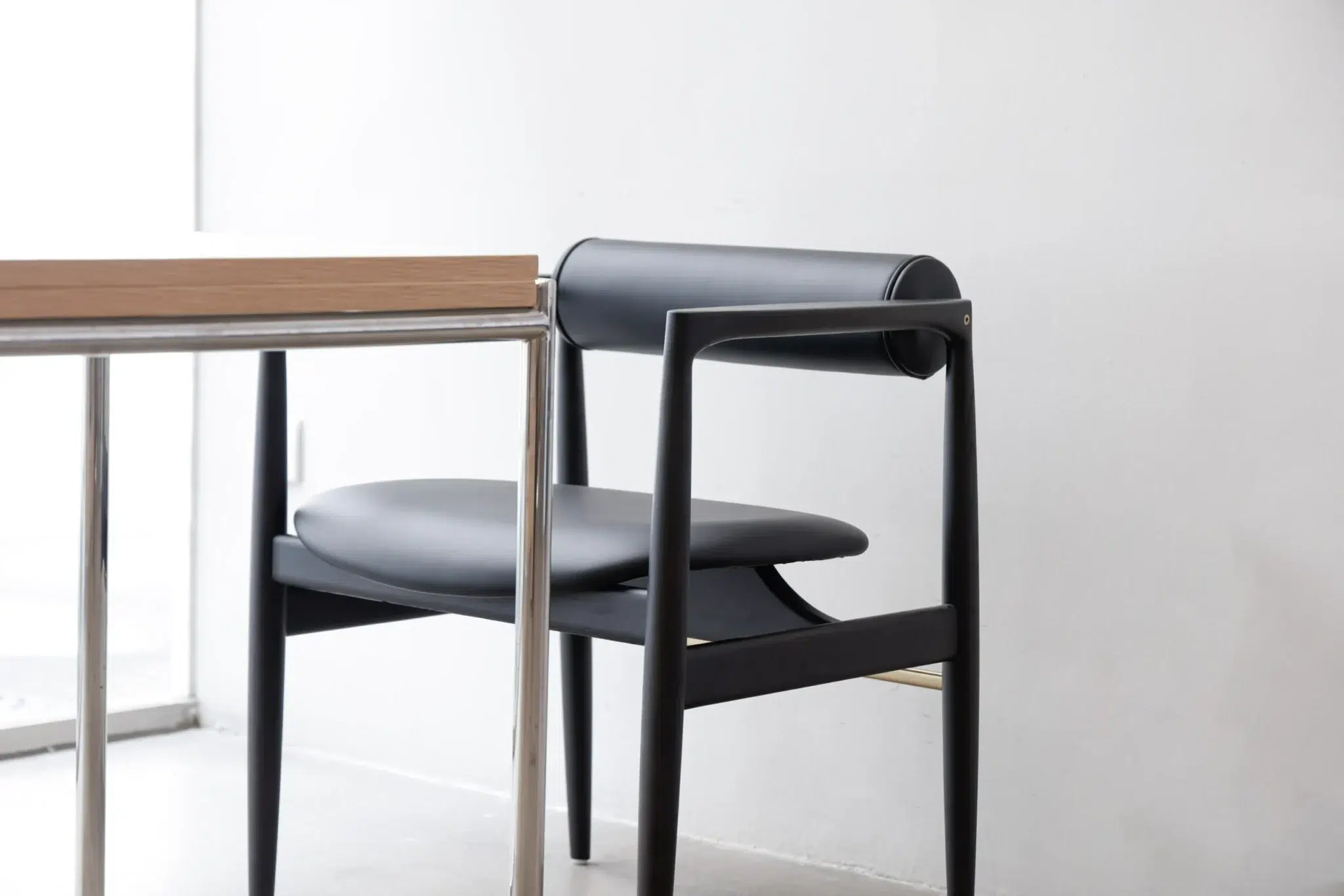 A modern black chair with a cylindrical backrest and a minimalistic design is placed next to a table with a light wooden top and metal legs in a bright, white room.