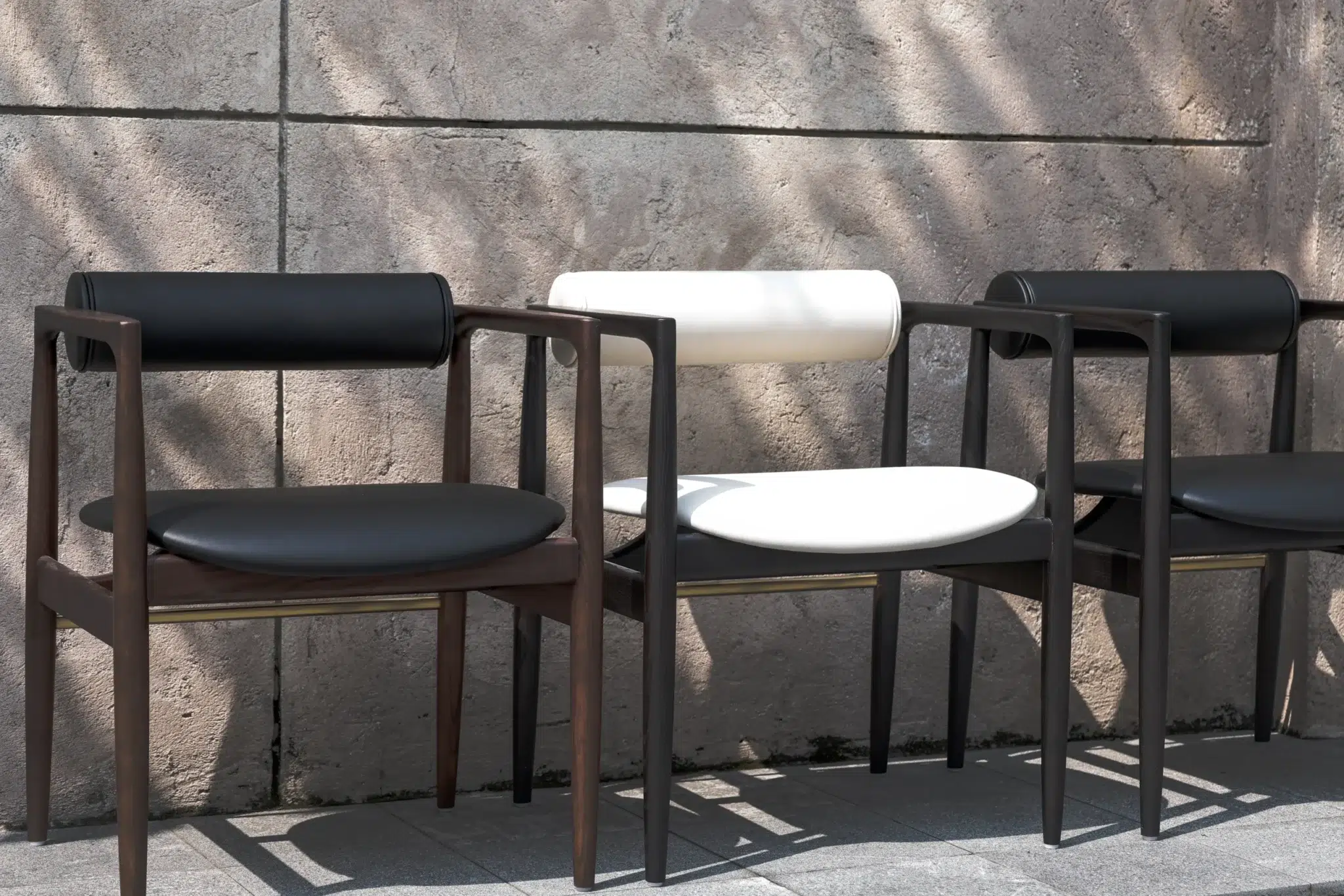 Three modern chairs with rounded seats and backs, featuring dark wooden frames. Two chairs are black, and one in the centre is white. They are placed outdoors against a textured concrete wall with dappled sunlight.