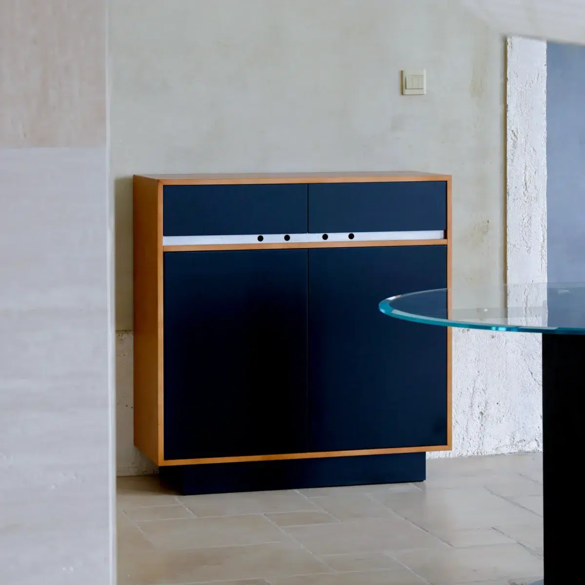 Elly Cabinet 2.0 A modern cupboard with a wooden frame, dark blue doors and drawers, and a white accent strip stands against a light-coloured wall in a minimalist room with tiled floors and a glass-topped table nearby.
