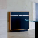 A modern cupboard with a wooden frame, dark blue doors and drawers, and a white accent strip stands against a light-coloured wall in a minimalist room with tiled floors and a glass-topped table nearby.