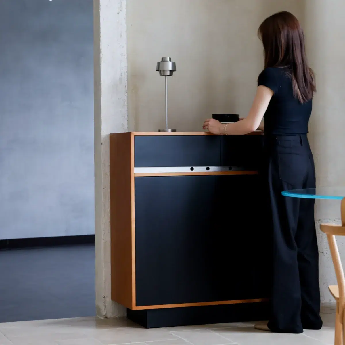 Elly Cabinet 2.0 A woman with long hair, wearing a black top and black trousers, stands in front of a modern black and wood cabinet, interacting with an object on top, beside a small table and chair in a minimalist room.