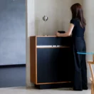 A woman with long hair, wearing a black top and black trousers, stands in front of a modern black and wood cabinet, interacting with an object on top, beside a small table and chair in a minimalist room.