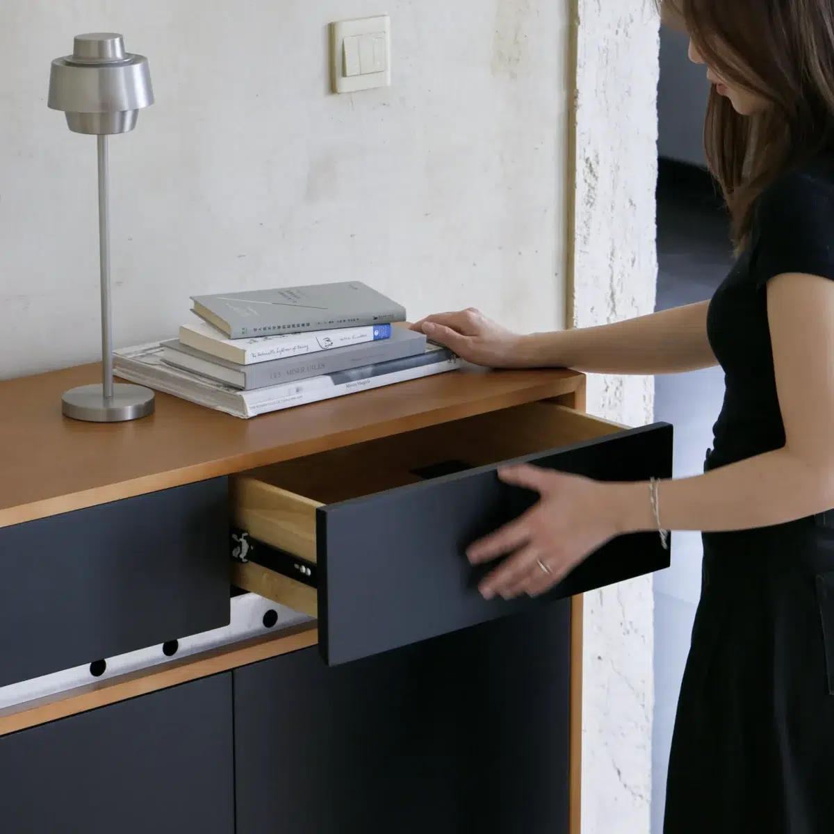 Elly Cabinet 2.0 A woman in a black dress opens a black drawer in a modern cupboard with a wooden top. A stack of books and a silver lamp rest on the cupboard surface against a light-coloured wall.